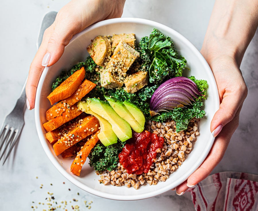 Woman hands eating vegan salad of baked vegetables, avocado, tofu and buckwheat buddha bowl, top view. Plant based food concept.