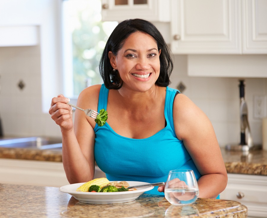 Smiling lady holding a folk with broccoli on it, with a plate of healthy food on the table